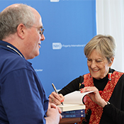 Dr. Jody Olsen, wearing a black sweater incorporating an orange pattern, signs a copy of her memoir for a man seen from behind and wearing a blue shirt.